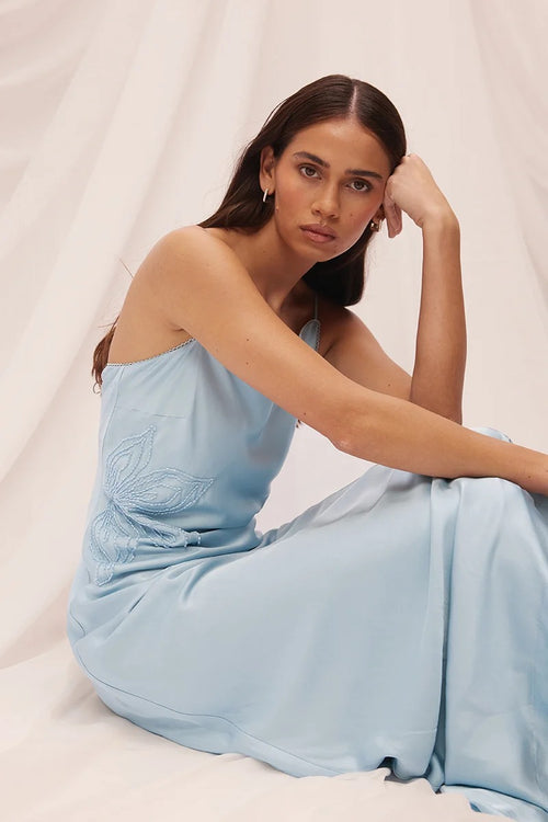 A woman wearing a light blue satin slip dress with delicate embroidery on the bodice, posing in a studio setting with draped white fabric.