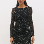 A woman modeling a black long-sleeve mini dress with white polka dots and sheer mesh sleeves, standing against a plain light gray studio background.