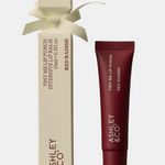 Ashley & Co Red Radish Intensive Lip Balm shown with its cream-colored box tied in a ribbon and the matching dark red lip balm tube on a light gray background.