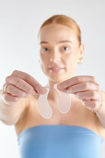 A woman holds up two translucent, crescent-shaped silicone eye patches in front of her face against a light gray background.