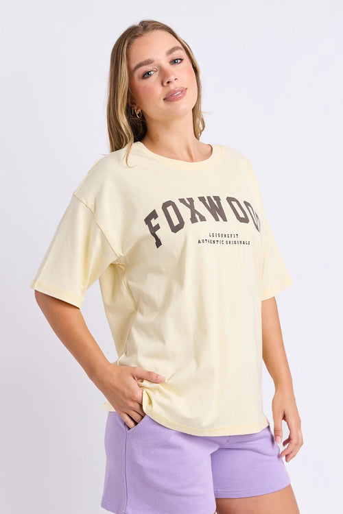 A woman modeling a pale yellow Foxwood short-sleeve t-shirt with dark brown collegiate lettering, paired with lavender shorts, against a light gray studio background.