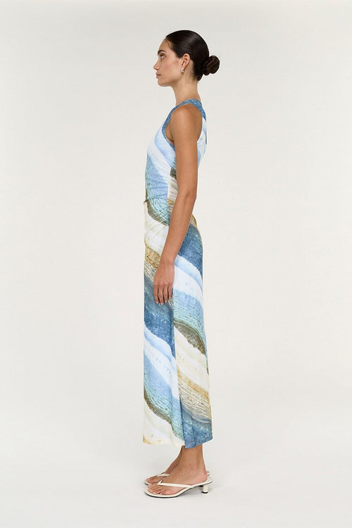 A woman in profile wearing a sleeveless maxi dress with a blue, beige, and white abstract pattern, paired with white heeled sandals, against a plain light gray studio background.