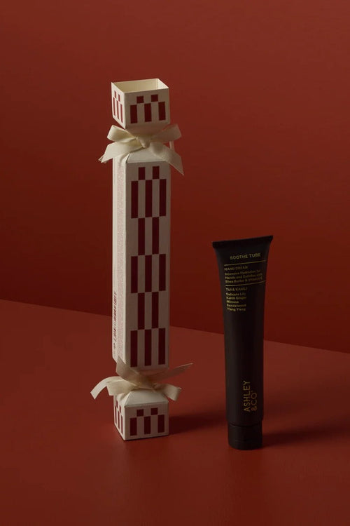 A cosmetic product set on a warm red studio background, featuring a tall cream-colored box with red vertical stripes and white ribbon bows next to a black squeeze tube with gold text.