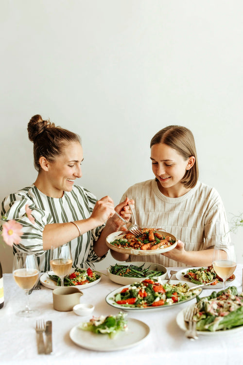 Two women sitting at a dining table sharing a meal with plates of salad and a main dish, in a bright, minimalist room.