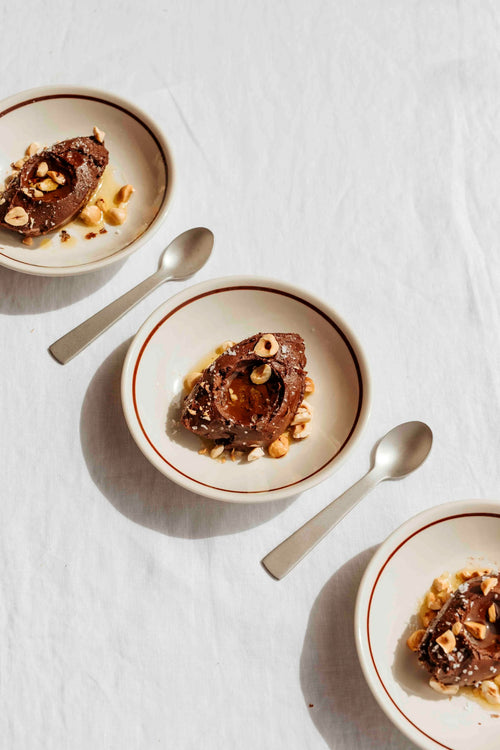 Three ceramic bowls with a thin red stripe border, each containing a chocolate dessert topped with chopped nuts, arranged on a white textured fabric with silver spoons.