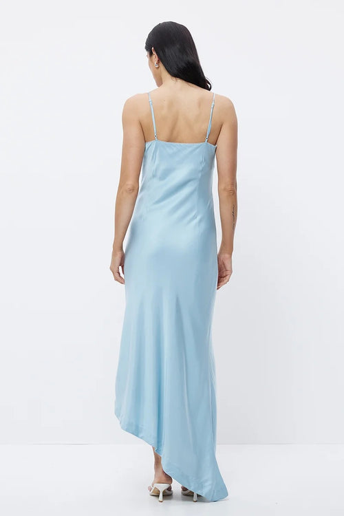 A woman viewed from the back wearing a light blue satin slip dress with thin spaghetti straps and an asymmetrical hem, paired with white heeled sandals, against a plain white studio background.