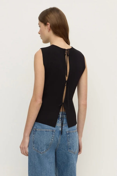 A woman shown from the back wearing a black sleeveless top with a tie-back detail and blue jeans, photographed against a plain light background.