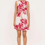 A woman modeling a sleeveless white A-line dress with a large pink and red floral print, paired with clear high-heeled sandals, set against a plain white studio background.