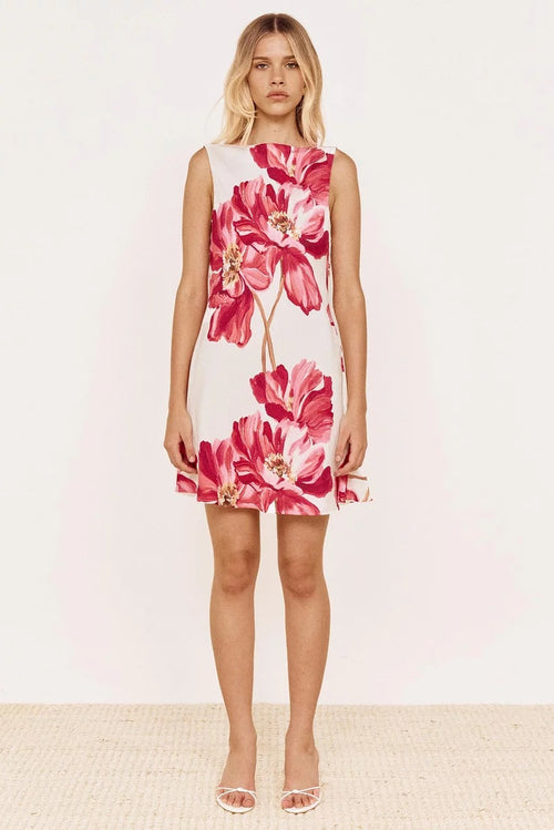 A woman modeling a sleeveless white A-line dress with a large pink and red floral print, paired with clear high-heeled sandals, set against a plain white studio background.