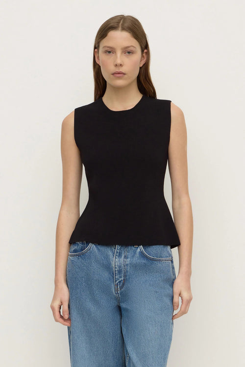 A woman modeling a sleeveless black knit top with a peplum waist, paired with blue jeans, against a plain off-white background.