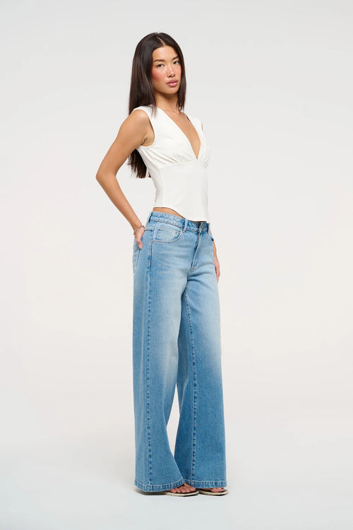 Model wearing mid indigo wide-leg jeans with a mid-rise waist, styled with a white sleeveless V-neck top and sandals.