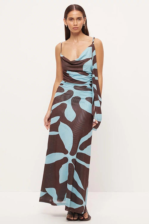 A woman modeling a sleeveless maxi dress with a brown and light blue abstract floral pattern, photographed against a plain white studio background.
