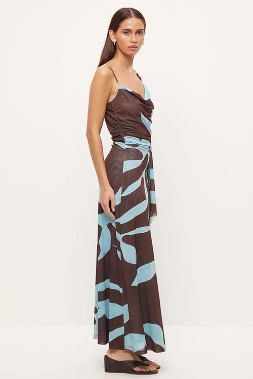 A woman modeling a brown maxi dress with a light blue abstract pattern and a cowl neckline, standing against a plain white studio background.