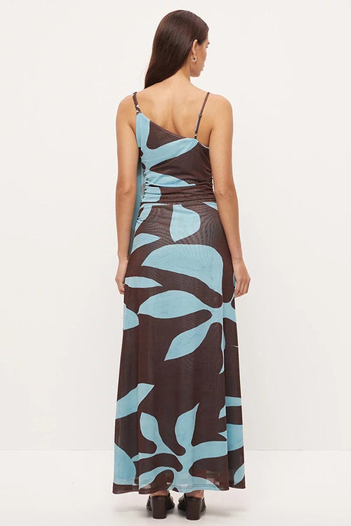 A woman viewed from the back wearing a sleeveless maxi dress with thin straps, a gathered waist, and an abstract leaf pattern in light blue and dark brown on a white studio background.