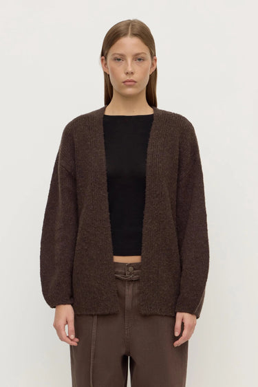 Person wearing a brown cardigan over a black top and brown pants on a white background