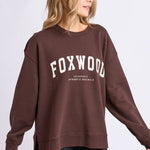 Woman wearing a brown sweatshirt with 'FOXWOOD' printed on it against a light gray background