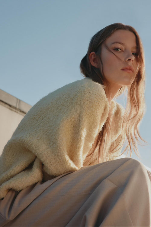 A woman wearing a cream-colored fuzzy knit sweater and beige wide-leg trousers, photographed from a low angle against a clear blue sky.