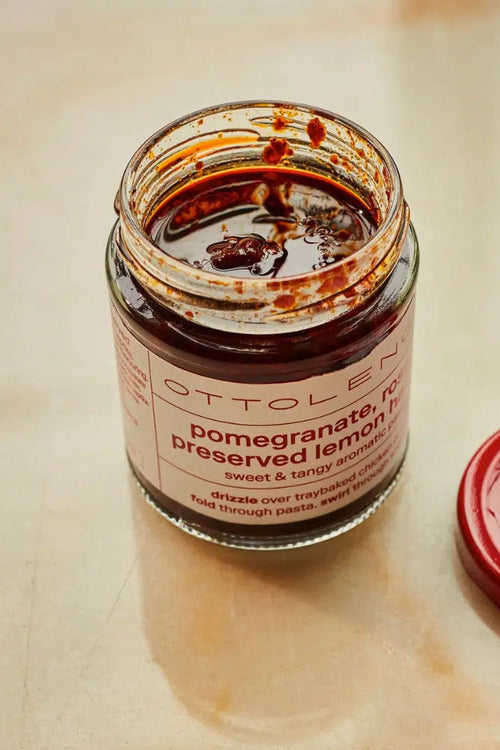 An open jar of Ottolen pomegranate preserved lemon harissa with its red lid placed beside it on a light beige surface. The jar contains a dark reddish-brown paste, and the label reads 'OTTOLEN pomegranate, no preserved lemon harissa sweet & tangy aduki'.