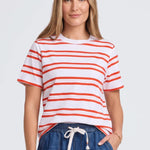A woman modeling a white short-sleeve crewneck t-shirt with thin orange horizontal stripes, paired with blue denim drawstring shorts, against a light gray studio background.