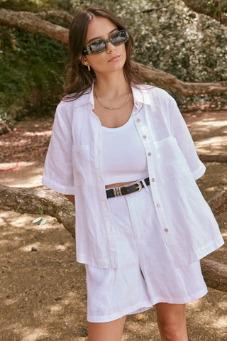 model wears a matching white button up top and shorts set with a white singlet underneath and black glasses