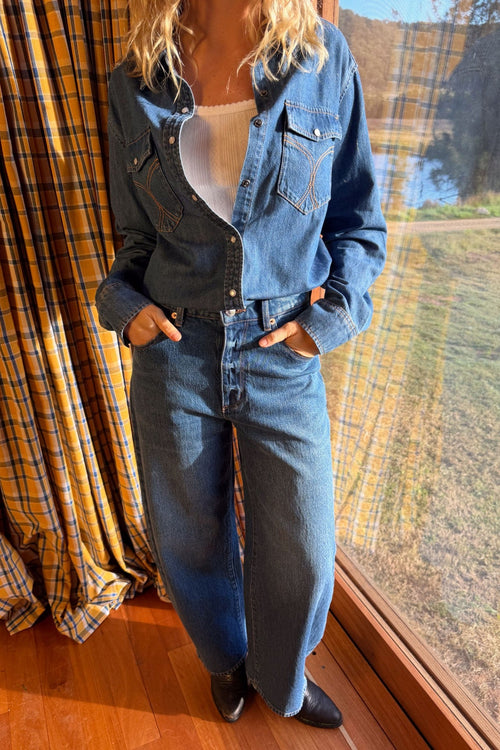 A woman wearing a cropped blue denim shirt-jacket and matching wide-leg denim jeans, standing indoors near a large window with a view of grass and trees outside. She has her hands in her jeans pockets and is wearing black boots.