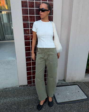 A woman stands outdoors wearing a white ribbed short-sleeve top, olive green wide-leg pants, black flats, and a white fuzzy shoulder bag, posing in front of a building with red-brown tiled columns.