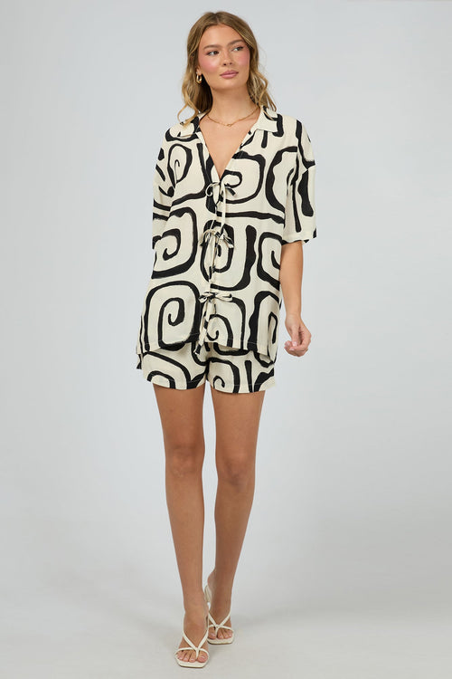 cream buttonless shirt with bold black swirl print and multiple front tie fastenings, worn with gold jewellery