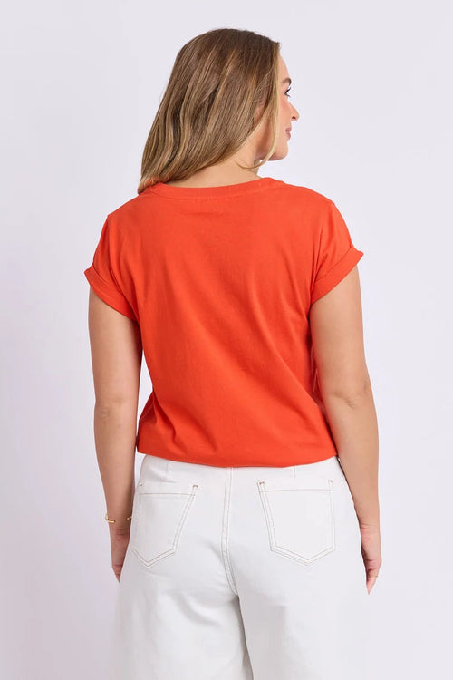 Woman wearing an orange t-shirt and white pants on a light purple background