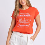 A woman modeling a bright orange short-sleeve graphic t-shirt with white text and sailboat illustrations reading 'French Riviera' and 'Foxwood', paired with white pants, against a light gray studio background.