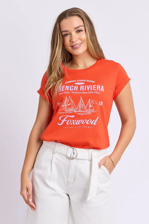 A woman modeling a bright orange short-sleeve graphic t-shirt with white text and sailboat illustrations reading 'French Riviera' and 'Foxwood', paired with white pants, against a light gray studio background.
