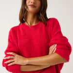 A woman wearing a fuzzy bright pink crewneck sweater and leopard-print pants, posing with her arms crossed against a plain light gray background.
