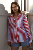 Woman wearing a red and white striped shirt and denim shorts standing against a white wall.